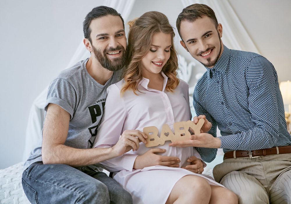 Gay couple becoming parents through surrogacy. An expectant mom sitting on the bed, loving fathers-to-be at her sides. The men smiling happily while holding lettering BABY over the pregnant belly.